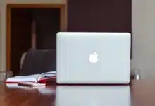 An Apple brand Macbook sits on a brown desk next to an open notebook.
