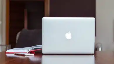 An Apple brand Macbook sits on a brown desk next to an open notebook.