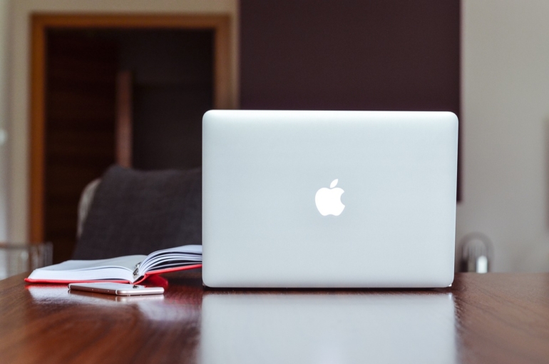 Una Macbook de la marca Apple descansa sobre un escritorio marrón junto a una libreta abierta.