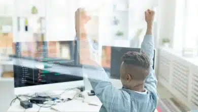 Ecstatic young African-American software developer raising hands in excitement while achieving success in web development, he sitting in front of computer behind glassy wall